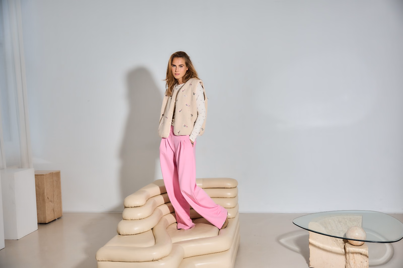 Model in pinker Hose und Strickweste steht auf einem cremefarbenen Sofa